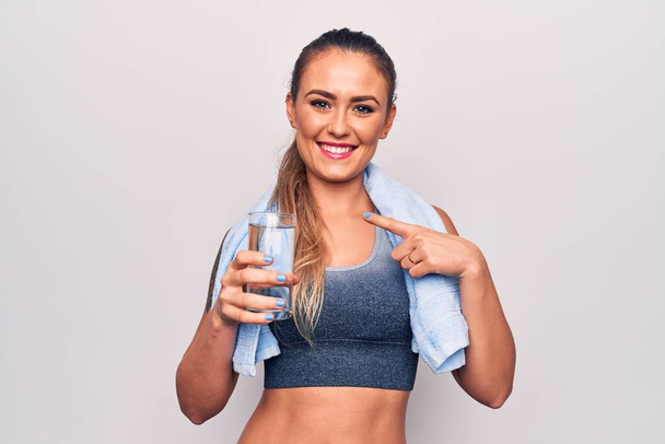 Bella donna bionda sportiva utilizzando asciugamano bottiglia di acqua su sfondo bianco sorridente felice puntando con mano e dito - Foto, immagini
