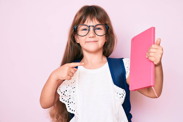 Petite fille enfant caucasienne aux cheveux longs portant un sac à dos étudiant et tenant un livre souriant heureux pointant avec la main et le doigt  - Photo, image