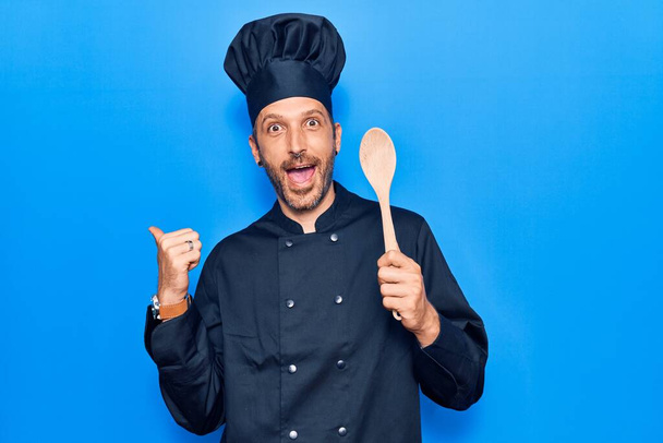 Joven hombre guapo con uniforme de cocina sosteniendo cuchara apuntando el pulgar hacia el lado sonriendo feliz con la boca abierta  - Foto, Imagen