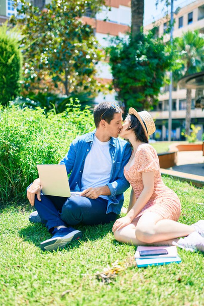 Beautiful couple kissing sitting on the grass using laptop at park - Photo, Image