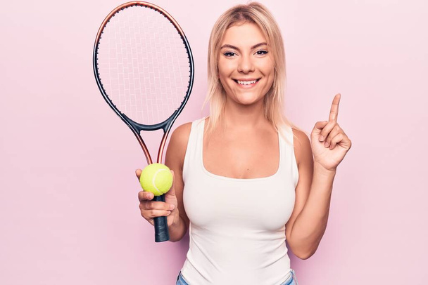 Młoda piękna blond sportowiec gra w tenisa za pomocą rakiety i piłkę na różowym tle uśmiechając się z pomysłem lub pytanie wskazując palec ze szczęśliwą twarzą, numer jeden - Zdjęcie, obraz