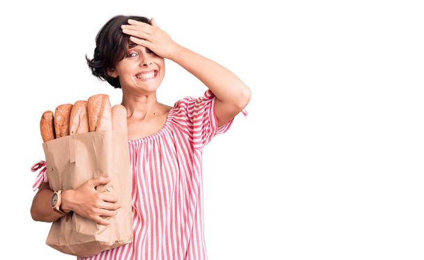Beautiful young woman with short hair holding paper bag with bread stressed and frustrated with hand on head, surprised and angry face  - Foto, Imagen