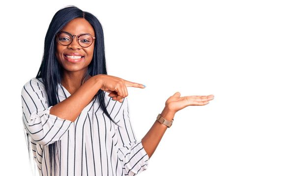 手で提示し、指で指している間にカジュアルな服や眼鏡を身に着けている若いアフリカ系アメリカ人の女性は、カメラに驚いて笑顔.  - 写真・画像