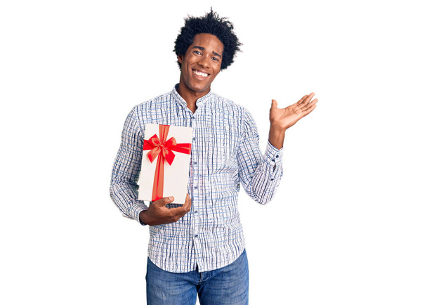 Bell'uomo afro-americano con capelli afro che tiene in mano il regalo celebrando la vittoria con sorriso felice e espressione del vincitore con le mani alzate  - Foto, immagini