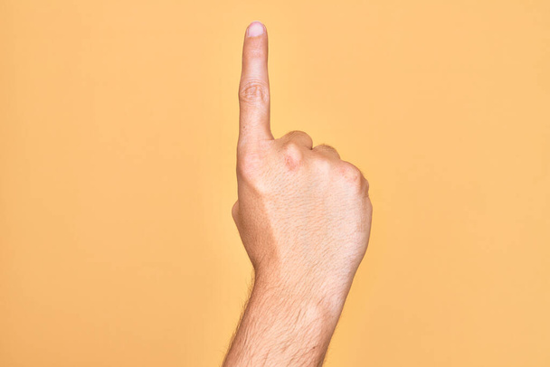 Hand eines kaukasischen jungen Mannes mit Fingern über isoliertem gelben Hintergrund, der die Zahl eins mit dem Zeigefinger zählt und dabei Idee und Verständnis zeigt - Foto, Bild