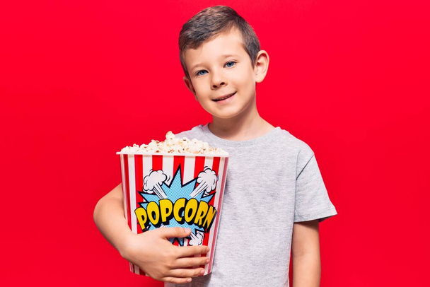 Roztomilé blonďaté dítě držící popcorn vypadá pozitivně a šťastně stojící a usmívá se s sebevědomým úsměvem ukazující zuby  - Fotografie, Obrázek