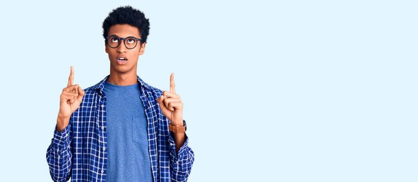 若いアフリカ系アメリカ人の男性は、カジュアルな服や眼鏡を身に着けている指と腕を上げて見て驚いています.  - 写真・画像