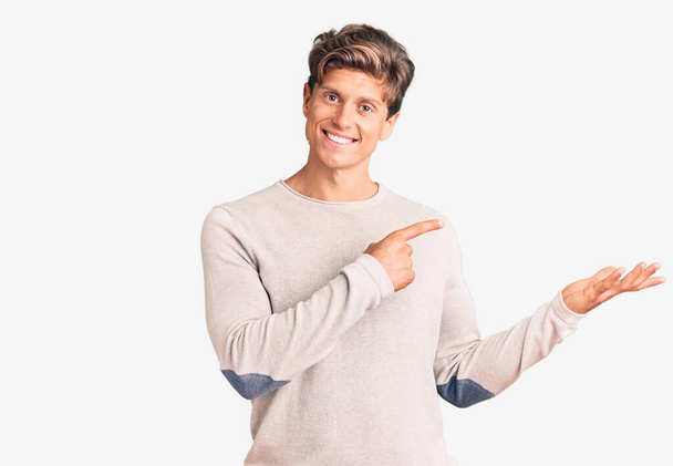 Junger gutaussehender Mann in lässiger Kleidung staunt und lächelt in die Kamera, während er mit der Hand präsentiert und mit dem Finger zeigt.  - Foto, Bild