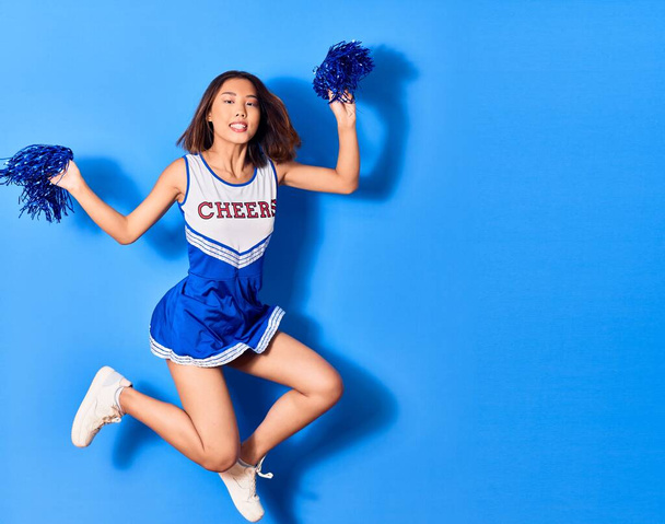 Giovane bella ragazza cinese sorridente felice indossa uniforme cheerleader. Salto con sorriso sul viso utilizzando pompom su sfondo blu isolato - Foto, immagini
