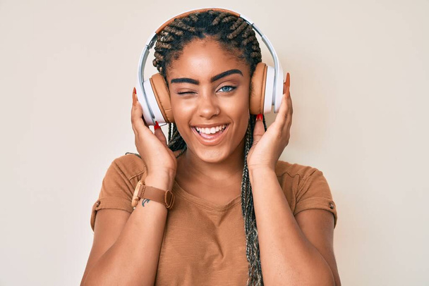 Saçları örgülü, kulaklıkla müzik dinleyen, kameraya seksi bir ifadeyle bakan, neşeli ve mutlu yüzlü genç Afro-Amerikan kadın..  - Fotoğraf, Görsel