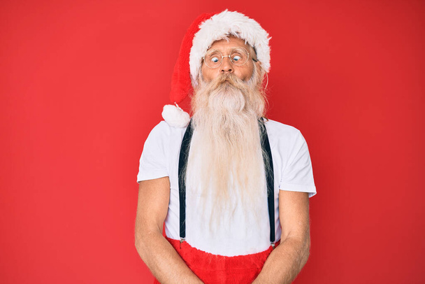 Γέρος ηλικιωμένος άνδρας με γκρίζα μαλλιά και μακριά γενειάδα φορώντας λευκό t-shirt και κοστούμι Santa Claus κάνοντας ψαρομούρη με χείλη, τρελή και κωμική χειρονομία. αστεία έκφραση.  - Φωτογραφία, εικόνα