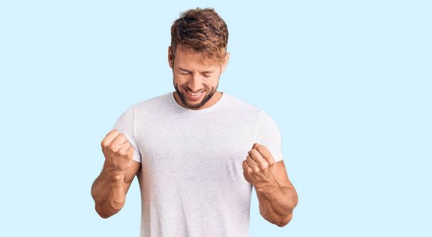 Jeune homme caucasien portant un t-shirt blanc décontracté très heureux et excité faisant geste gagnant avec les bras levés, souriant et criant pour le succès. concept de célébration.  - Photo, image