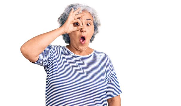 Oudere vrouw met grijs haar draagt casual gestreepte kleren doet ok gebaar geschokt met verrast gezicht, oog kijken door vingers. ongelovige uitdrukking.  - Foto, afbeelding