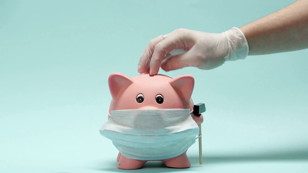 Ausgeschnittene Ansicht eines Mannes in Latex-Handschuhen, der Geld in Sparschwein auf blau legt  - Filmmaterial, Video