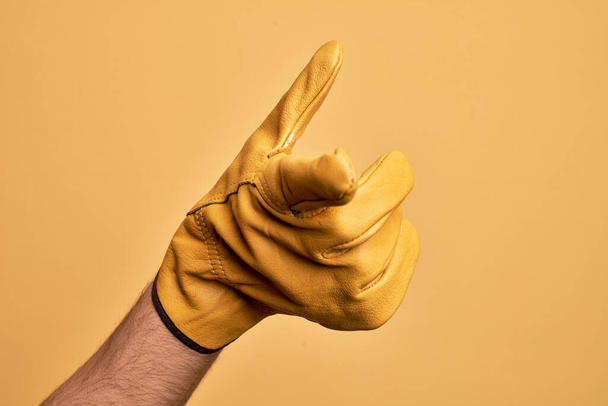 Χέρι του Καυκάσου νεαρός άνδρας με γάντι κηπουρού πάνω από απομονωμένο κίτρινο φόντο δείχνει δείκτη προς την κάμερα, επιλέγοντας και υποδεικνύοντας προς την κατεύθυνση - Φωτογραφία, εικόνα