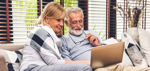 一緒にラップトップコンピュータを使用して楽しい時間を過ごしているシニア夫婦。幸せな高齢者の夫と妻は、ソーシャルメディアをチェックし、自宅でソファに座っている間にニュースやショッピングをオンラインで読んでいます。退職の概念 - 写真・画像