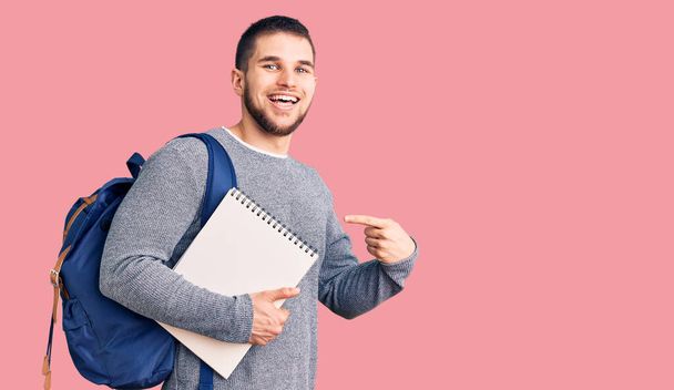 Joven hombre guapo con mochila de estudiante sosteniendo portátil sonriendo feliz señalando con la mano y el dedo  - Foto, Imagen