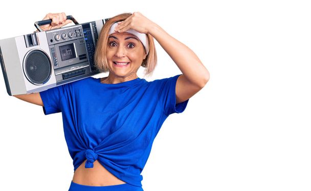 Young blonde woman wearing sportswear holding boombox, listening to music stressed and frustrated with hand on head, surprised and angry face  - Фото, изображение