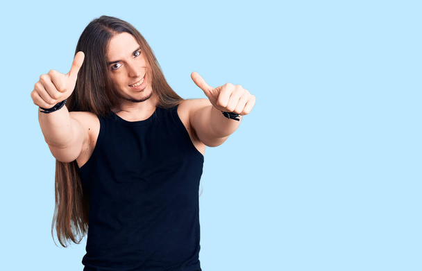 Young adult man with long hair wearing goth style with black clothes approving doing positive gesture with hand, thumbs up smiling and happy for success. winner gesture.  - Fotoğraf, Görsel