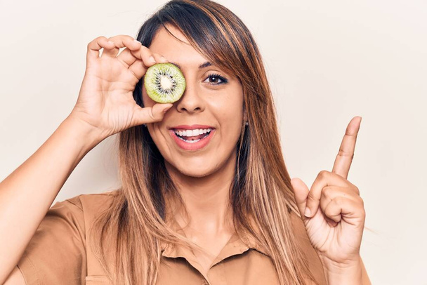 Young beautiful woman holding kiwi over eye smiling happy pointing with hand and finger to the side  - 写真・画像
