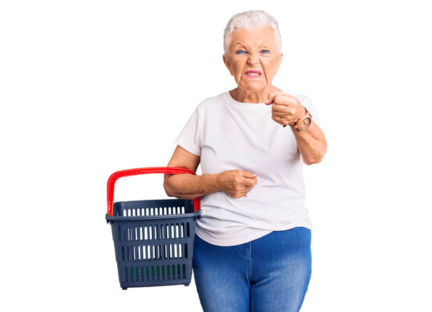 Senior kaunis nainen siniset silmät ja harmaat hiukset tilalla supermarket ostoskori harmitti ja turhautunut huutaa vihaa, huutaa hullu vihaa ja käsi esille  - Valokuva, kuva