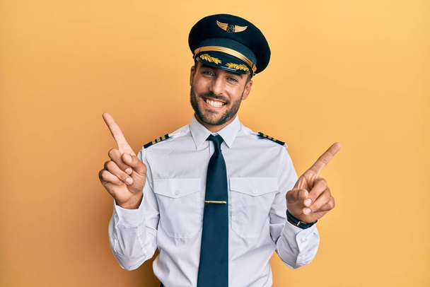Przystojny Hiszpan w mundurze pilota samolotu, uśmiechający się z przekonaniem wskazując palcami w różnych kierunkach. miejsce na reklamę  - Zdjęcie, obraz