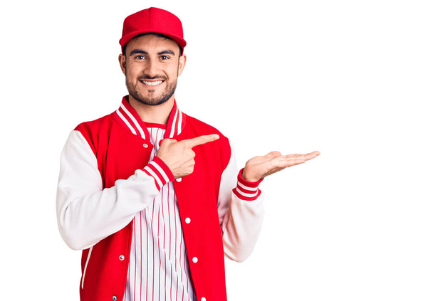 Joven hombre guapo con chaqueta de béisbol y gorra asombrado y sonriendo a la cámara mientras se presenta con la mano y señalando con el dedo.  - Foto, imagen