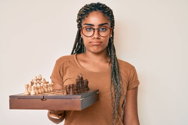 Молода афроамериканська жінка з плечима, що тримає шахову дошку в окулярах скептично і нервово, насуплена через проблеми. негативна людина.  - Фото, зображення