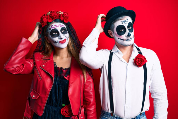 Couple wearing day of the dead costume over red confuse and wondering about question. uncertain with doubt, thinking with hand on head. pensive concept.  - Photo, Image
