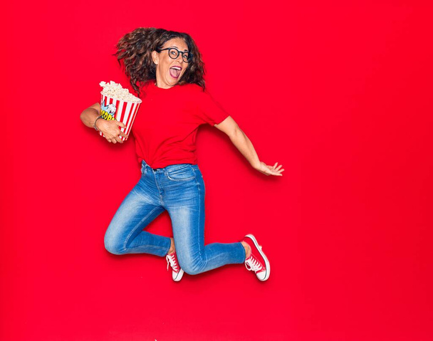 Middle age beautiful hispanic woman surprised with open mouth wearing glasses. Holding bucket of popcorn jumping over isolated red background - Fotografie, Obrázek