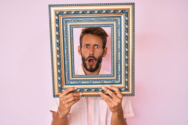 Handsome caucasian man with beard holding empty frame in shock face, looking skeptical and sarcastic, surprised with open mouth  - Photo, Image