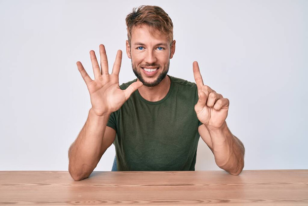 Jonge blanke man draagt casual kleding zittend op de tafel tonen en wijzen met vingers nummer zes, terwijl glimlachen zelfverzekerd en gelukkig.  - Foto, afbeelding
