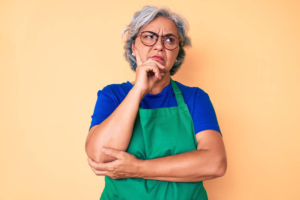 Ältere hispanische Frau mit Schürze und Brille ernstes Gesicht über Frage nachdenkend mit der Hand am Kinn, nachdenklich über verwirrende Idee  - Foto, Bild