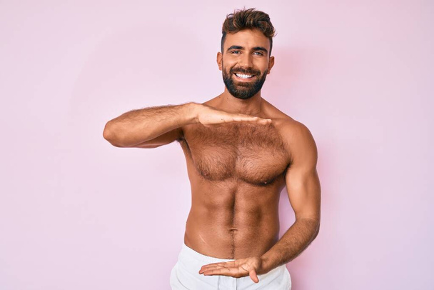 Young hispanic man standing shirtless gesturing with hands showing big and large size sign, measure symbol. smiling looking at the camera. measuring concept.  - Foto, Imagem