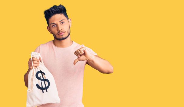 Handsome latin american young man holding money bag with dollar symbol with angry face, negative sign showing dislike with thumbs down, rejection concept  - Φωτογραφία, εικόνα