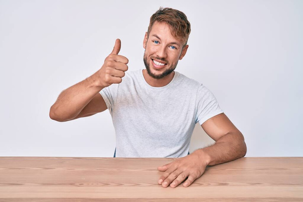 Mladý kavkazský muž v neformálním oblečení sedí na stole a usmívá se šťastně a pozitivně, palec nahoru dělá vynikající a schvalovací znamení  - Fotografie, Obrázek