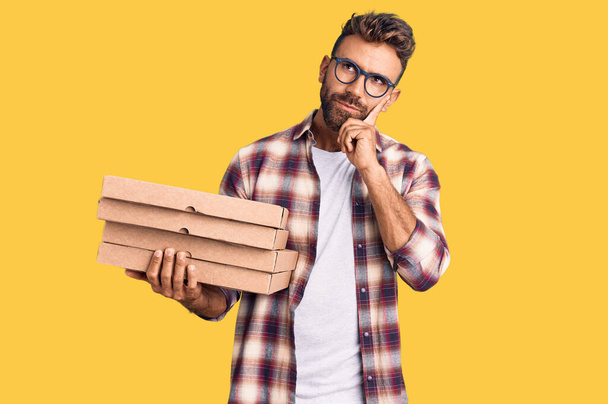 Young hispanic man holding delivery pizza box serious face thinking about question with hand on chin, thoughtful about confusing idea  - Foto, Bild