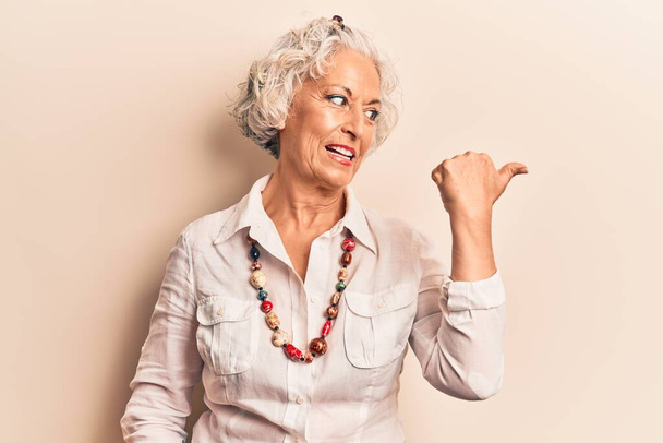 Senior grey-haired woman wearing casual clothes smiling with happy face looking and pointing to the side with thumb up.  - Photo, image