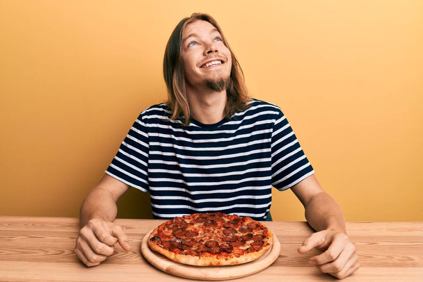 Schöner kaukasischer Mann mit langen Haaren, der leckere Pfefferoni-Pizza isst und mit einem Lächeln im Gesicht und natürlichem Gesichtsausdruck zur Seite schaut. Lachen selbstbewusst.  - Foto, Bild
