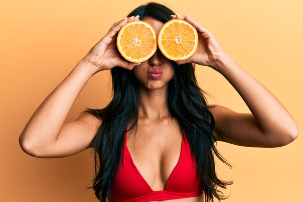 Piękna Latynoska kobieta w bikini trzymająca pomarańczowe oczy nadęte policzki z zabawną twarzą. usta napompowane powietrzem, łapie powietrze.  - Zdjęcie, obraz