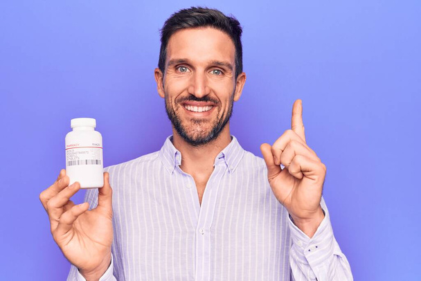 Молодой красивый мужчина держит банку с лекарствами, стоящую на изолированном фиолетовом фоне, улыбаясь, указывая рукой и пальцем в сторону. - Фото, изображение
