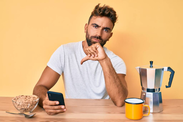 テーブルの上に座っている若いヒスパニック系の男は、朝食を持っていて、怒っている顔をしたスマートフォンを使用して、否定的なサインは親指で嫌いを示す、拒否の概念  - 写真・画像