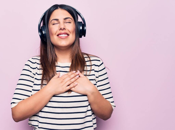 Mladá krásná brunetka, která poslouchá hudbu pomocí sluchátek na růžovém pozadí, usmívá se s rukama na hrudi, oči zavřené vděčným gestem na tváři. Zdravotní koncepce. - Fotografie, Obrázek