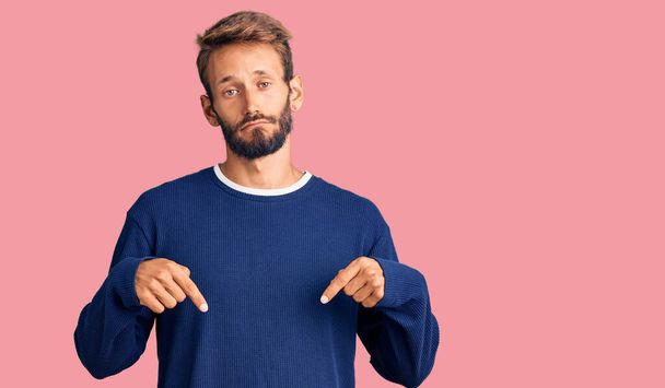 Hombre rubio guapo con barba usando suéter casual apuntando hacia abajo buscando triste y molesto, indicando la dirección con los dedos, infeliz y deprimido.  - Foto, imagen