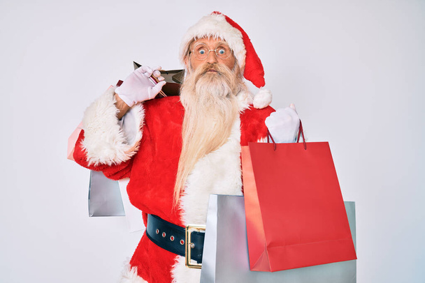 Oude man met grijs haar en lange baard in kerstman kostuum met boodschappentassen die wangen poetsen met een grappig gezicht. mond opgeblazen met lucht, vangen van lucht.  - Foto, afbeelding