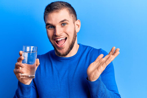 Genç yakışıklı adam bir bardak su içerek başarıyı mutlu bir gülümsemeyle ve kazanan yüz ifadesiyle kutluyor.  - Fotoğraf, Görsel