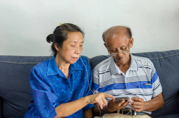 Ασιατικά ηλικιωμένα ζευγάρια παρακολουθούν κινητά τηλέφωνα και χρησιμοποιούν ακουστικά στον καναπέ. - Φωτογραφία, εικόνα