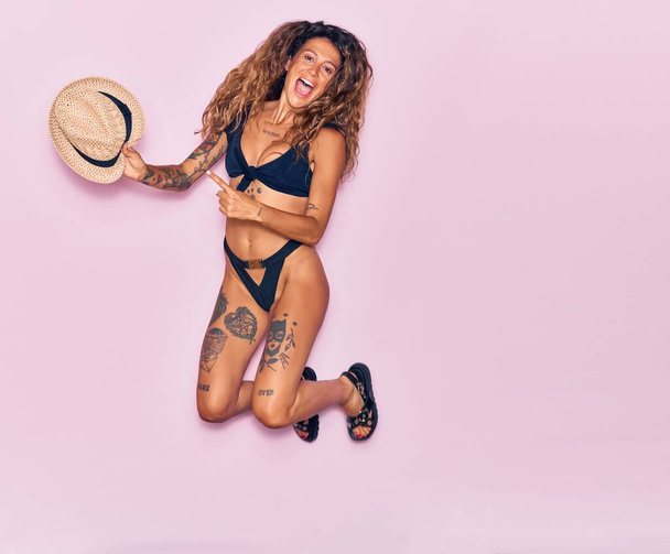 Young beautiful curly woman with tattoo on vacation wearing bikini smiling happy. Jumping with smile on face pointing finger to summer hat over isolated pink background. - Photo, Image