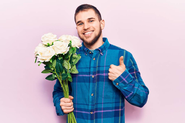 Jeune bel homme tenant bouquet de fleurs souriant heureux et positif, pouce levé faire excellent et signe d'approbation  - Photo, image