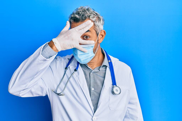 Середнього віку сірошерстий чоловік у формі лікаря і медична маска підглядає в шоковому покритті обличчя і очі рукою, дивлячись через пальці бояться
  - Фото, зображення